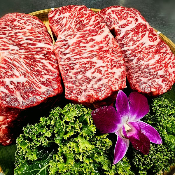 Three marbled wagyu beef steaks on a bamboo tray with green lettuce and a purple orchid.