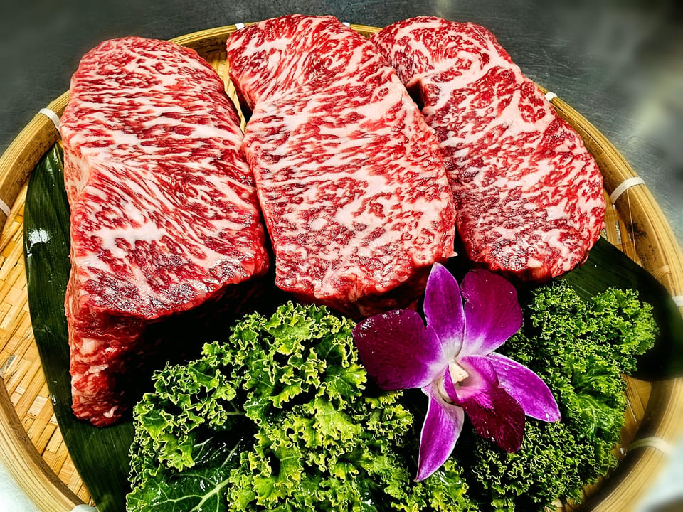 Three marbled wagyu beef steaks on a bamboo tray with green lettuce and a purple orchid.