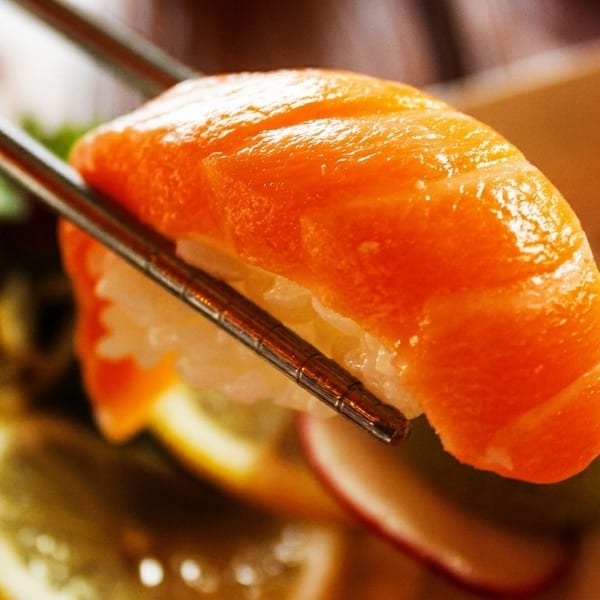 Chopsticks holding a piece of salmon nigiri sushi with rice, against a blurred background.